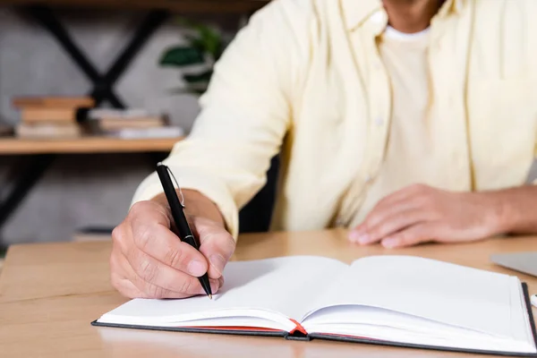 Vista parcial del hombre escribiendo en un cuaderno mientras trabajaba en casa, fondo borroso - foto de stock