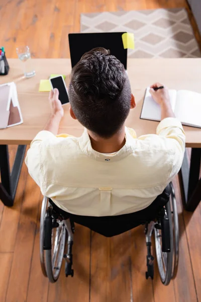 Visão aérea do homem com deficiência escrevendo no notebook enquanto trabalhava perto do laptop em casa — Fotografia de Stock