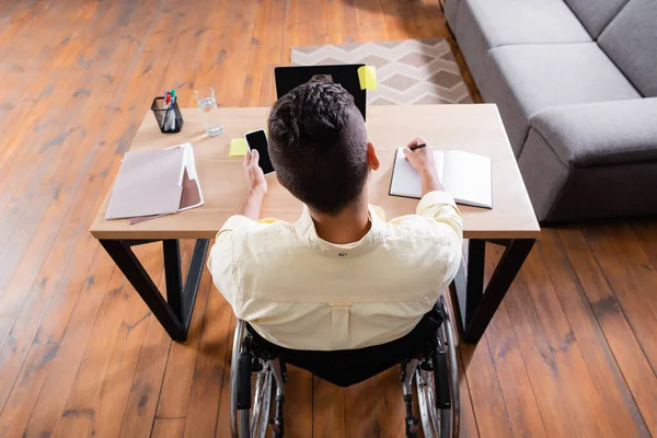 Blick auf Behinderten, der Handy in der Hand hält und in Notizbuch neben Laptop schreibt — Stockfoto