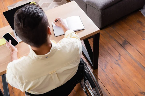 Overhead view of disabled man writing in notebook while holding smartphone with blank screen — Stock Photo