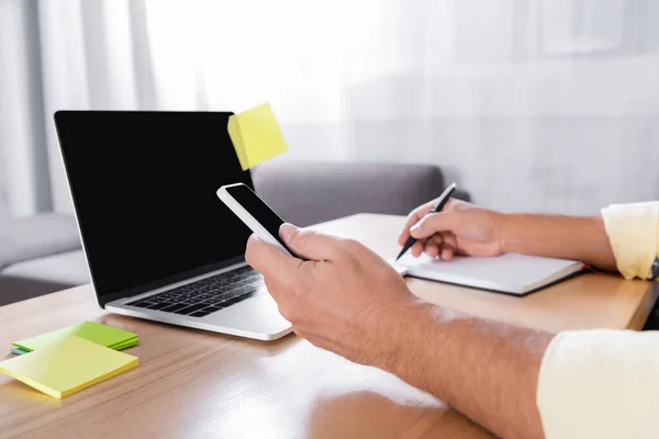 Cropped view of freelancer writing in notebook while holding smartphone near laptop with blank screen — Stock Photo