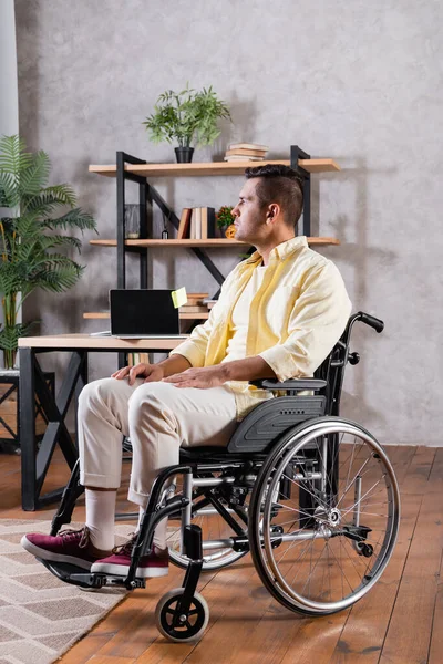 Hombre discapacitado en silla de ruedas mirando hacia otro lado cerca del ordenador portátil con pantalla en blanco en el escritorio - foto de stock