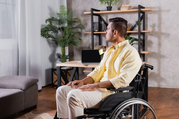 Deficiente homem sentado em cadeira de rodas perto de laptop com tela em branco na mesa — Stock Photo