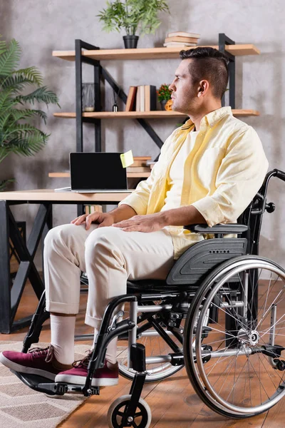 Hombre discapacitado mirando hacia otro lado mientras está sentado en silla de ruedas cerca de la computadora en el escritorio - foto de stock