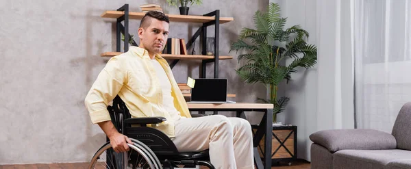 Инвалид смотрит в камеру, сидя в инвалидной коляске рядом с рабочим местом дома, баннер — стоковое фото