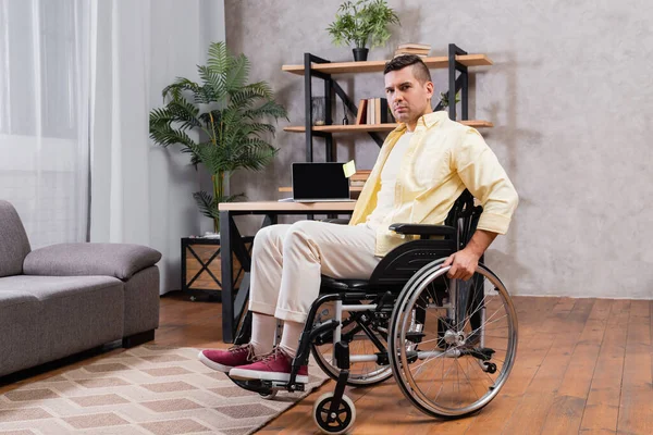 Молодий чоловік у інвалідному візку дивиться на камеру біля ноутбука з порожнім екраном на столі — стокове фото