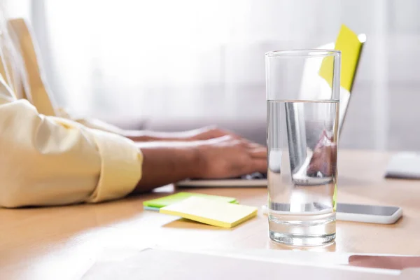 Foyer sélectif de verre avec de l'eau près de l'homme tapant sur ordinateur portable sur fond flou, vue recadrée — Photo de stock