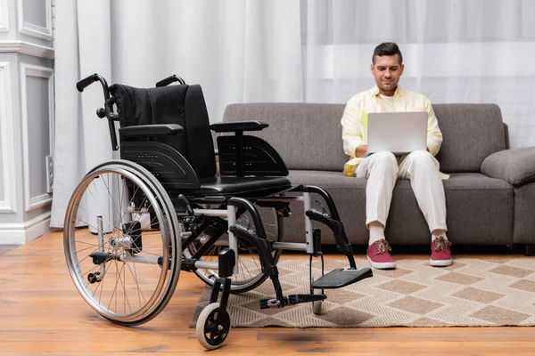 Homem com deficiência trabalhando no sofá com laptop perto de cadeira de rodas em primeiro plano — Fotografia de Stock