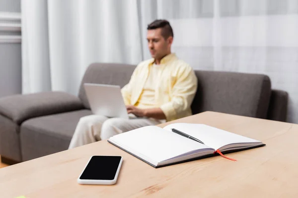 Selective focus of empty notebook and cellphone with blank screen near freelancer working on blurred background — Stock Photo