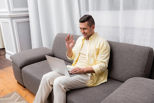 Hombre alegre saludando la mano durante la videollamada en el ordenador portátil en casa - foto de stock