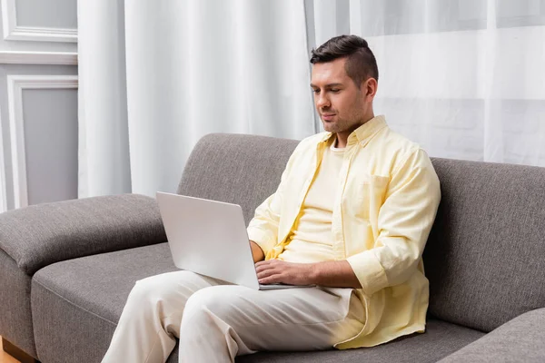 Молодий фрілансер в повсякденному одязі сидить на дивані з ноутбуком — стокове фото