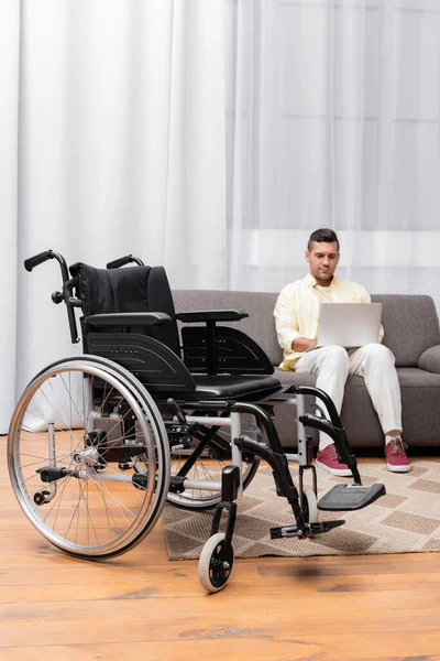 Handicapped man working on sofa with laptop near wheelchair on foreground — Stock Photo