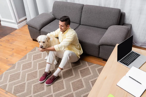 Visão de alto ângulo do homem acariciando cão labrador perto de laptop com tela em branco na mesa — Fotografia de Stock