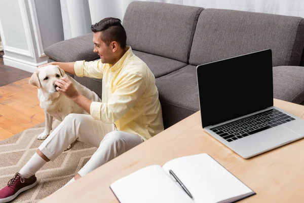 Hombre joven tocando la nariz del perro labrador mientras está sentado en el suelo cerca del escritorio con el ordenador portátil - foto de stock