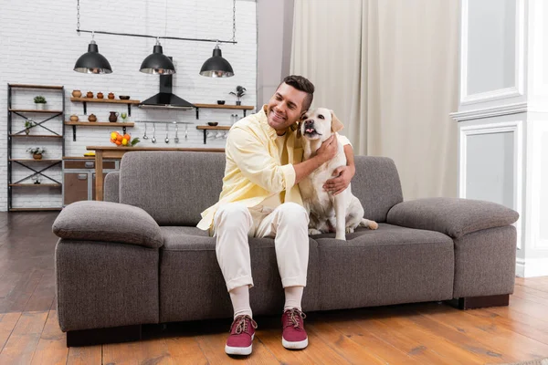 Glücklicher Mann mit geschlossenen Augen umarmt Labrador-Hund auf Couch zu Hause — Stockfoto