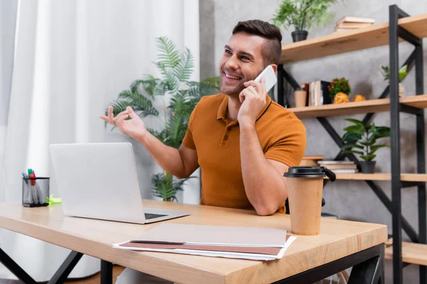 Fröhlicher, gehandicapter Mann gestikuliert, während er auf Smartphone neben Laptop spricht — Stockfoto