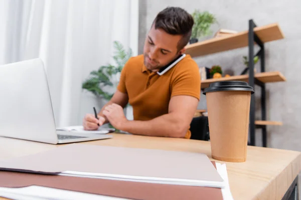 Enfoque selectivo de la taza de papel cerca freelancer hablando en el teléfono inteligente sobre fondo borroso - foto de stock