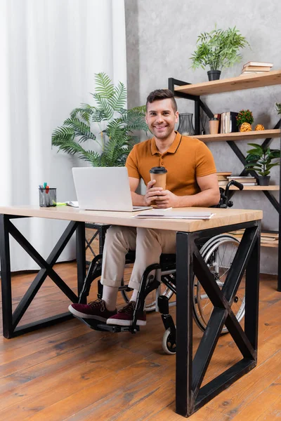 Uomo handicappato sorridente alla macchina fotografica mentre tiene il caffè per andare vicino al computer portatile — Foto stock