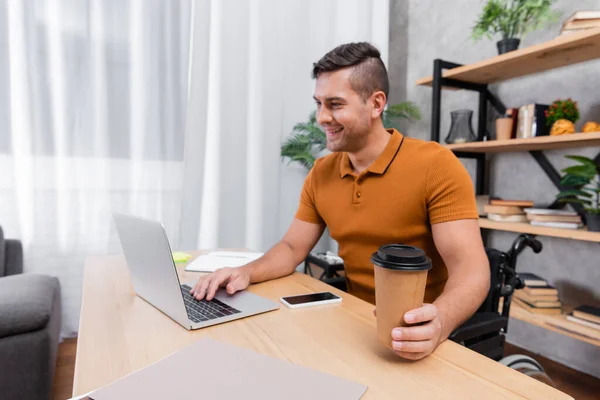 Positivo, homem com deficiência segurando café para ir enquanto digita no laptop perto do celular com tela em branco — Fotografia de Stock