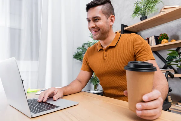 Alegre, homem deficiente segurando café para ir enquanto digita no laptop em casa — Fotografia de Stock