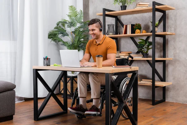 Sonriente, hombre discapacitado trabajando en auriculares cerca de la computadora portátil en la oficina en casa - foto de stock