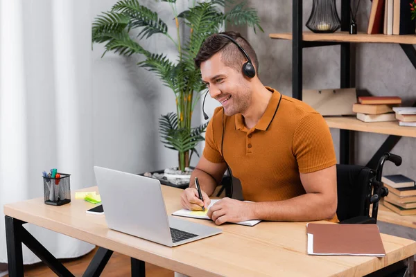 Lächelnder, behinderter Mann schreibt in Notizbuch, während er zu Hause im Headset arbeitet — Stockfoto