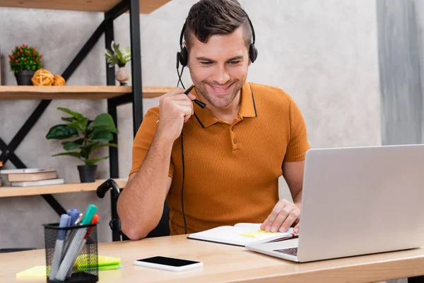Homem positivo, deficiente no fone de ouvido trabalhando perto de laptop e smartphone com tela em branco — Fotografia de Stock