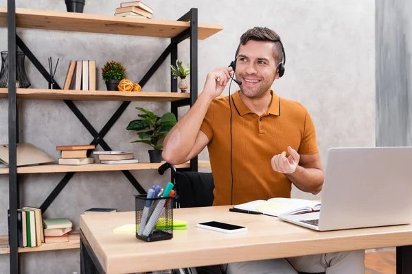 Homem alegre no fone de ouvido trabalhando em casa laptop puro e smartphone com tela em branco — Fotografia de Stock