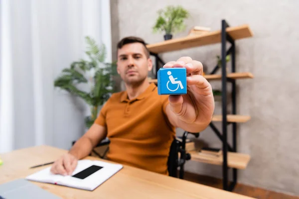 Jeune homme montrant un cube avec signe de handicap alors qu'il était assis au bureau à la maison, fond flou — Photo de stock