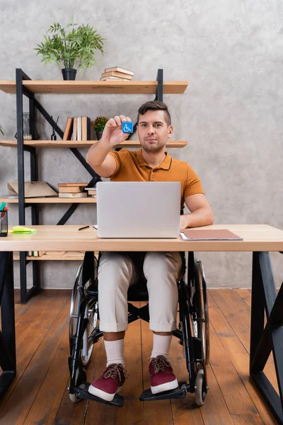 Junger Mann zeigt Behinderungszeichen, während er im Rollstuhl im Home Office sitzt — Stockfoto