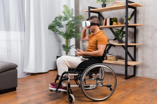 Uomo disabile sorridente mentre gioca in auricolare vr in sedia a rotelle a casa — Foto stock