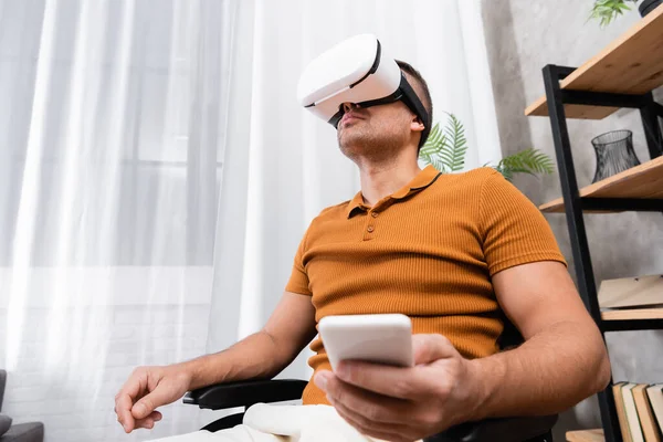 Low angle view of handicapped man holding smartphone while gaming in vr headset — Stock Photo