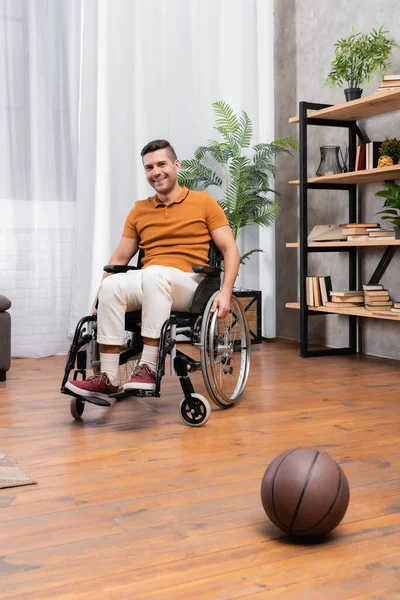 Позитивный инвалид сидит в инвалидной коляске рядом с баскетболом на полу — стоковое фото