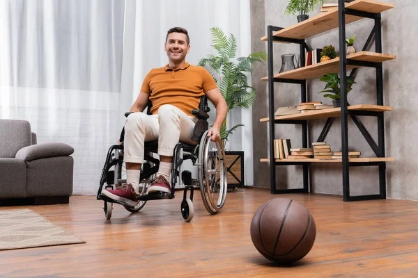 Fröhlicher Mann lächelt in die Kamera, während er im Rollstuhl neben Basketball auf dem Boden sitzt — Stockfoto
