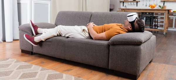Молодой человек в повседневной одежде, лежащий в гарнитуре на диване дома, баннер — стоковое фото
