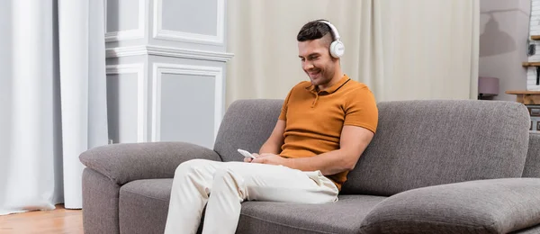 Homme souriant assis sur le canapé, bavarder sur le smartphone et écouter de la musique dans les écouteurs, bannière — Photo de stock