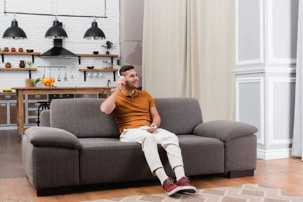 Uomo sorridente che regola le cuffie mentre ascolta musica sul divano di casa — Foto stock