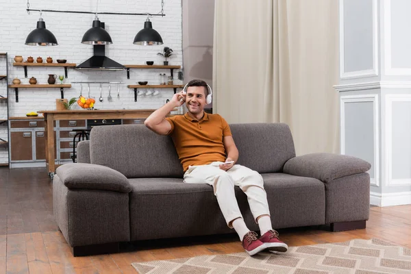 Fröhlicher Mann hört Musik über Kopfhörer, während er auf dem Sofa in einer modernen Wohnung sitzt — Stockfoto