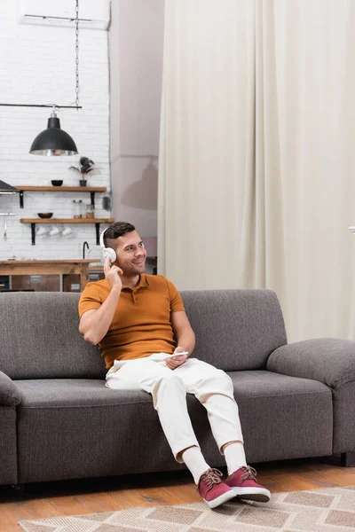 Усміхнений чоловік слухає музику в навушниках, сидячи на дивані зі смартфоном — стокове фото