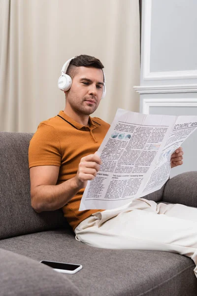 Young man reading newspaper and listening music in headphones near smartphone with blank screen — Stock Photo