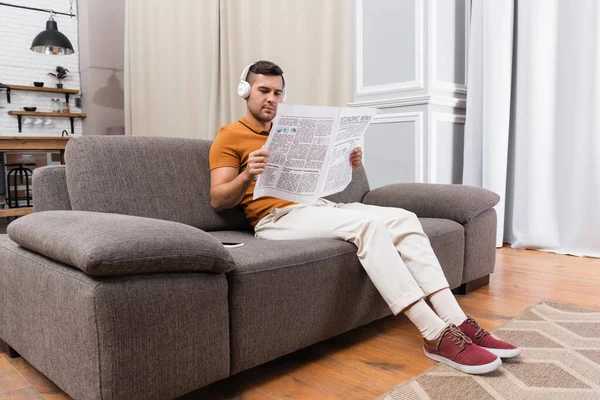 Hombre en auriculares escuchando música y leyendo periódicos en el sofá en casa - foto de stock