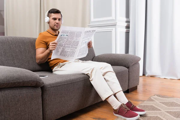 Young man reading newspaper while listening music in headphones on couch at home — Stock Photo