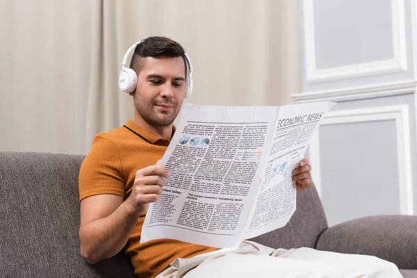 Hombre positivo en auriculares inalámbricos leyendo periódico en el sofá en casa - foto de stock