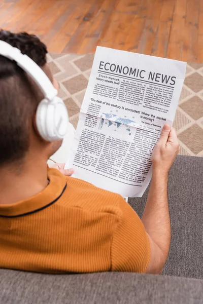 Vista aérea del hombre en auriculares inalámbricos que leen noticias económicas en primer plano borroso - foto de stock