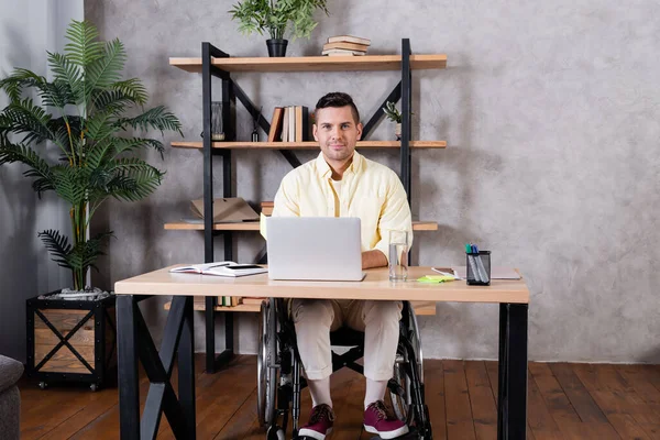 Hombre discapacitado mirando la cámara mientras está sentado en silla de ruedas cerca de la computadora portátil - foto de stock