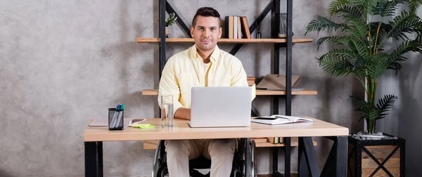 Homem com deficiência sorrindo para a câmera enquanto trabalhava perto do laptop em casa, banner — Fotografia de Stock