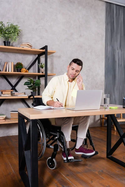 Disabled man writing in notebook and talking on mobile phone while working at home — Stock Photo