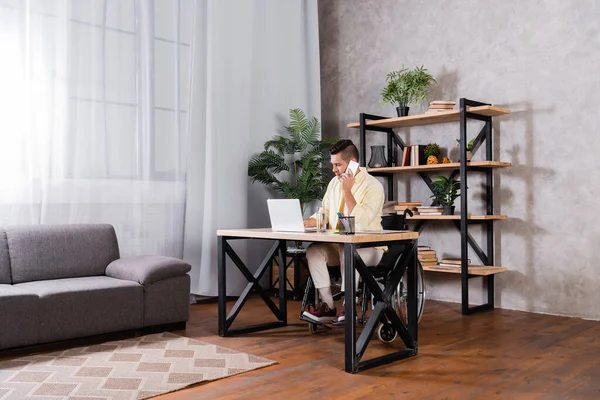 Disabled man talking on mobile phone while working near laptop in home office — Stock Photo