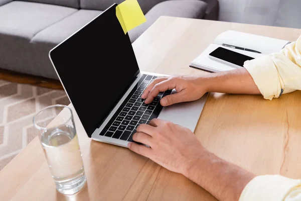 Partial view of freelancer typing on laptop with blank screen near glass of water — Stock Photo