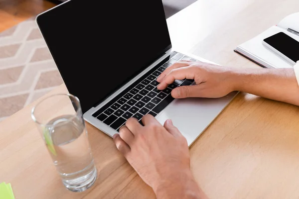 Partial view of man typing on laptop with blank screen near glass of water — Stock Photo